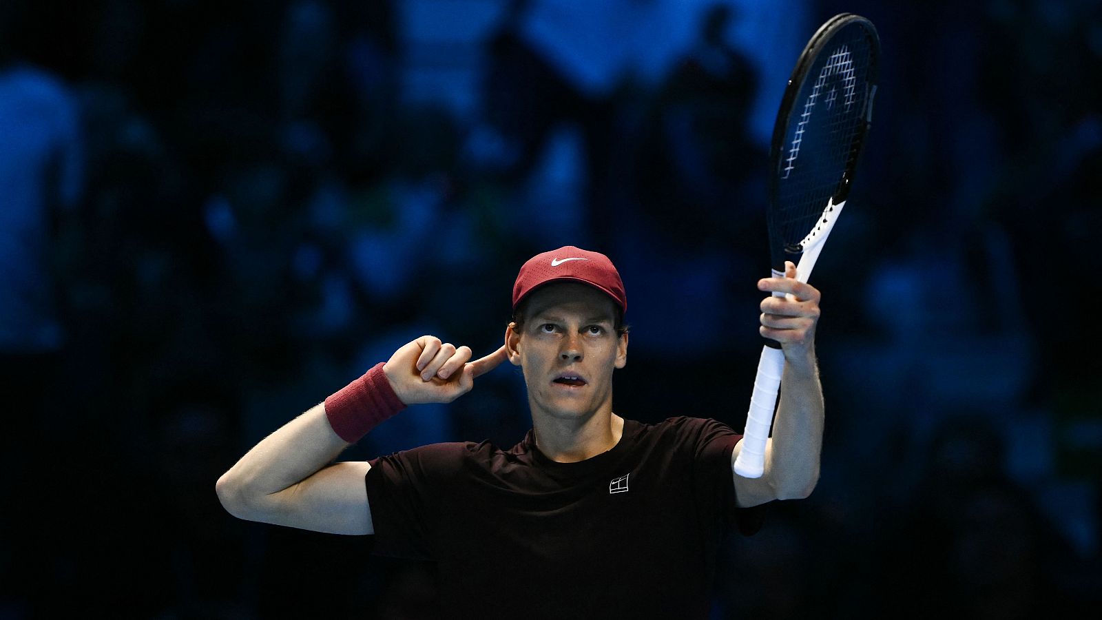 Jannik Sinner, con vestimenta deportiva, celebra un triunfo en una cancha de tenis, sosteniendo una raqueta y tocándose la oreja, con un público borroso de fondo.