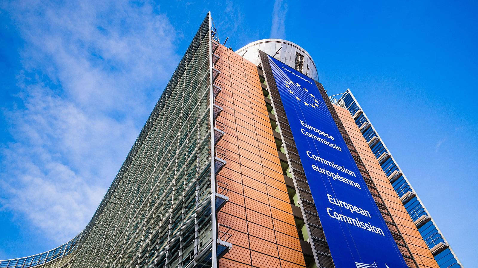 Un edificio moderno con fachada de cristal y metal, con una sección naranja y una estructura cilíndrica en la parte superior, exhibe una pancarta azul con el logo de la Unión Europea y texto multilingüe.