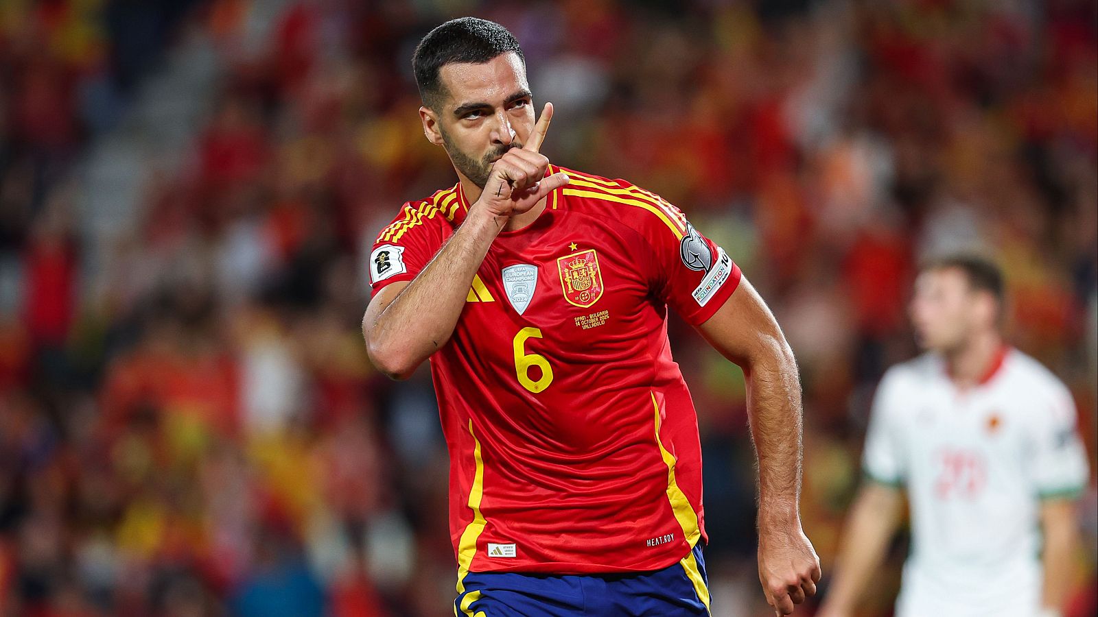 Un jugador de fútbol con la camiseta roja y el número 6, en un gesto de silencio, se encuentra en un campo con otros jugadores y público de fondo.