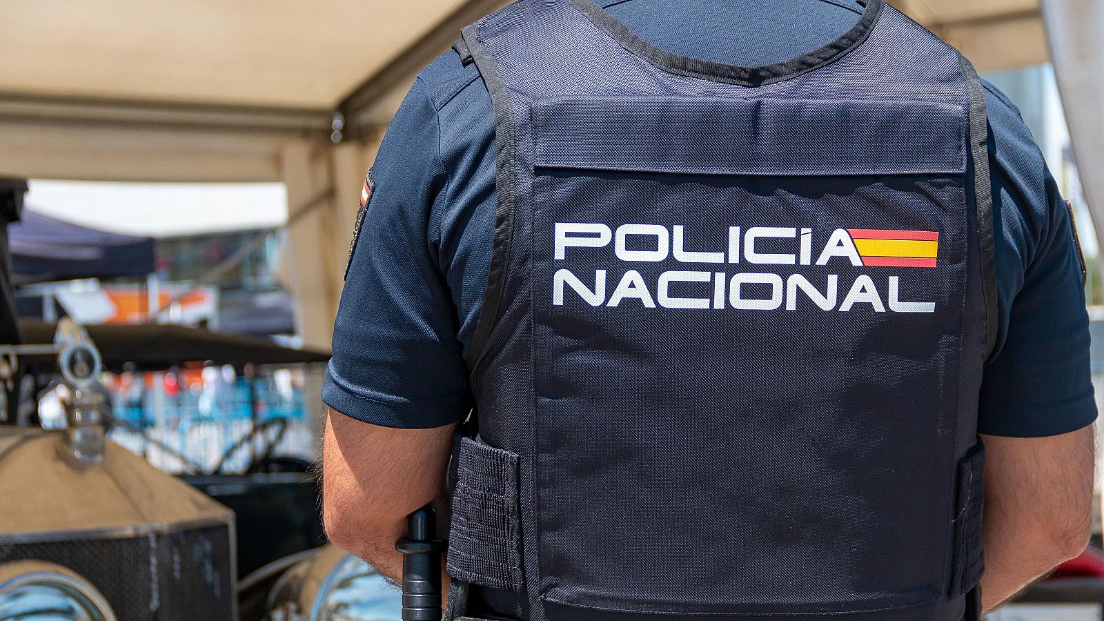 Un agente de la Policía Nacional, de espaldas, lleva un chaleco azul oscuro con la inscripción "POLICIA NACIONAL" y la bandera de España. Se observa parte de su uniforme y, al fondo, un coche y botellas.