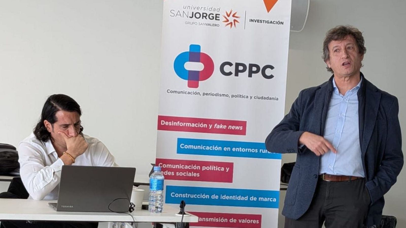 Dos hombres participan en una mesa redonda; uno gesticula y el otro escucha frente a un ordenador. Un cartel de la Universidad San Jorge con el logo y temas relacionados con la comunicación, periodismo y política, sirve de fondo.