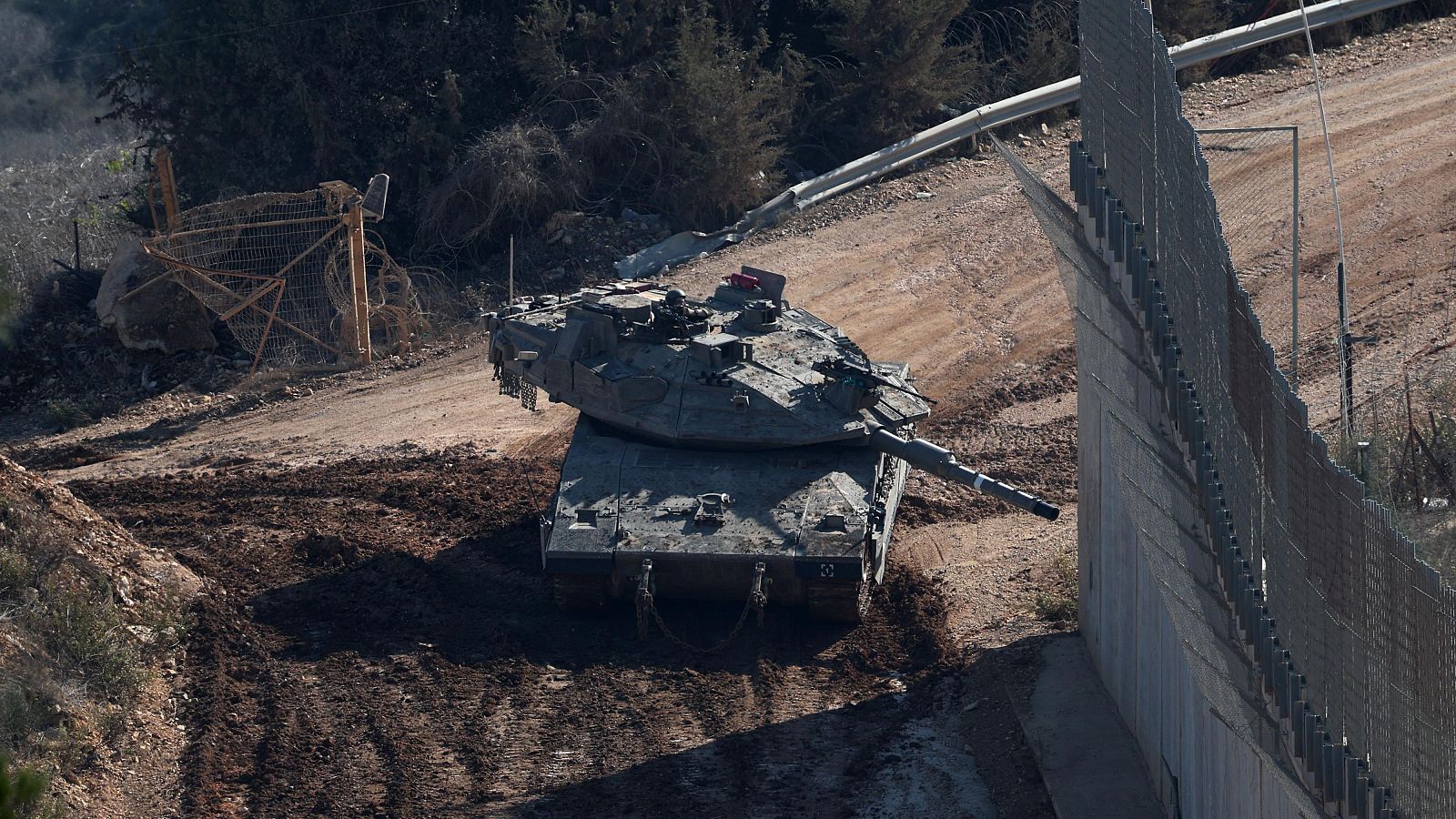Un tanque militar se sitúa cerca de una valla fronteriza en un entorno rocoso y con vegetación escasa, posiblemente en una zona de conflicto. Se observa una valla metálica dañada en el fondo.