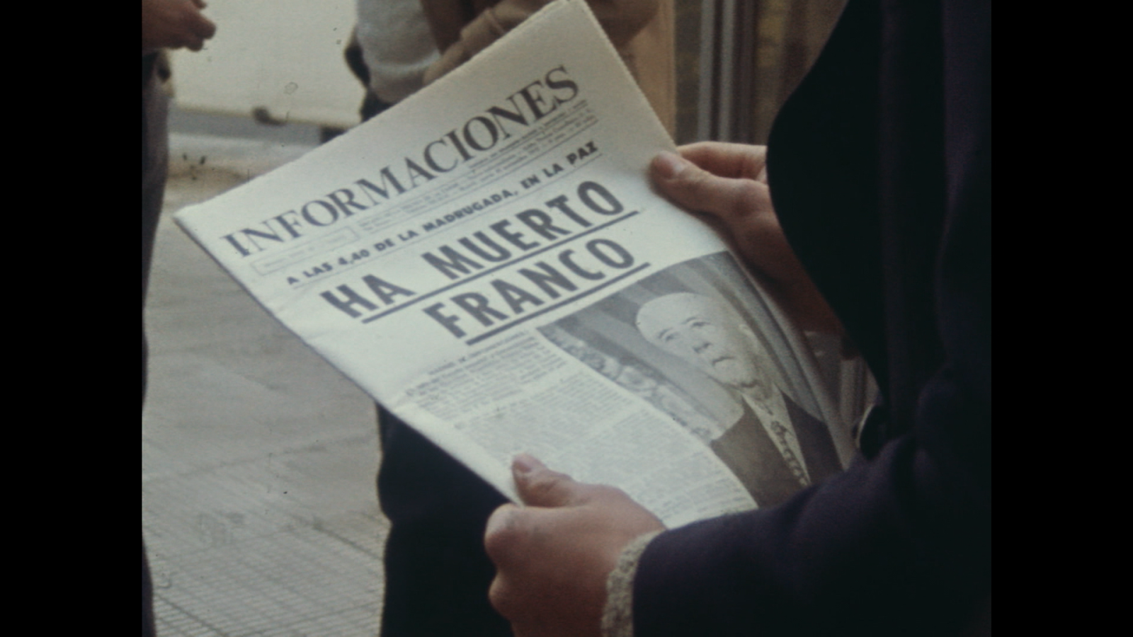 Una persona sostiene un periódico abierto con el titular que anuncia la muerte de un personaje relevante. En la parte inferior del periódico, se muestra una fotografía del mismo.