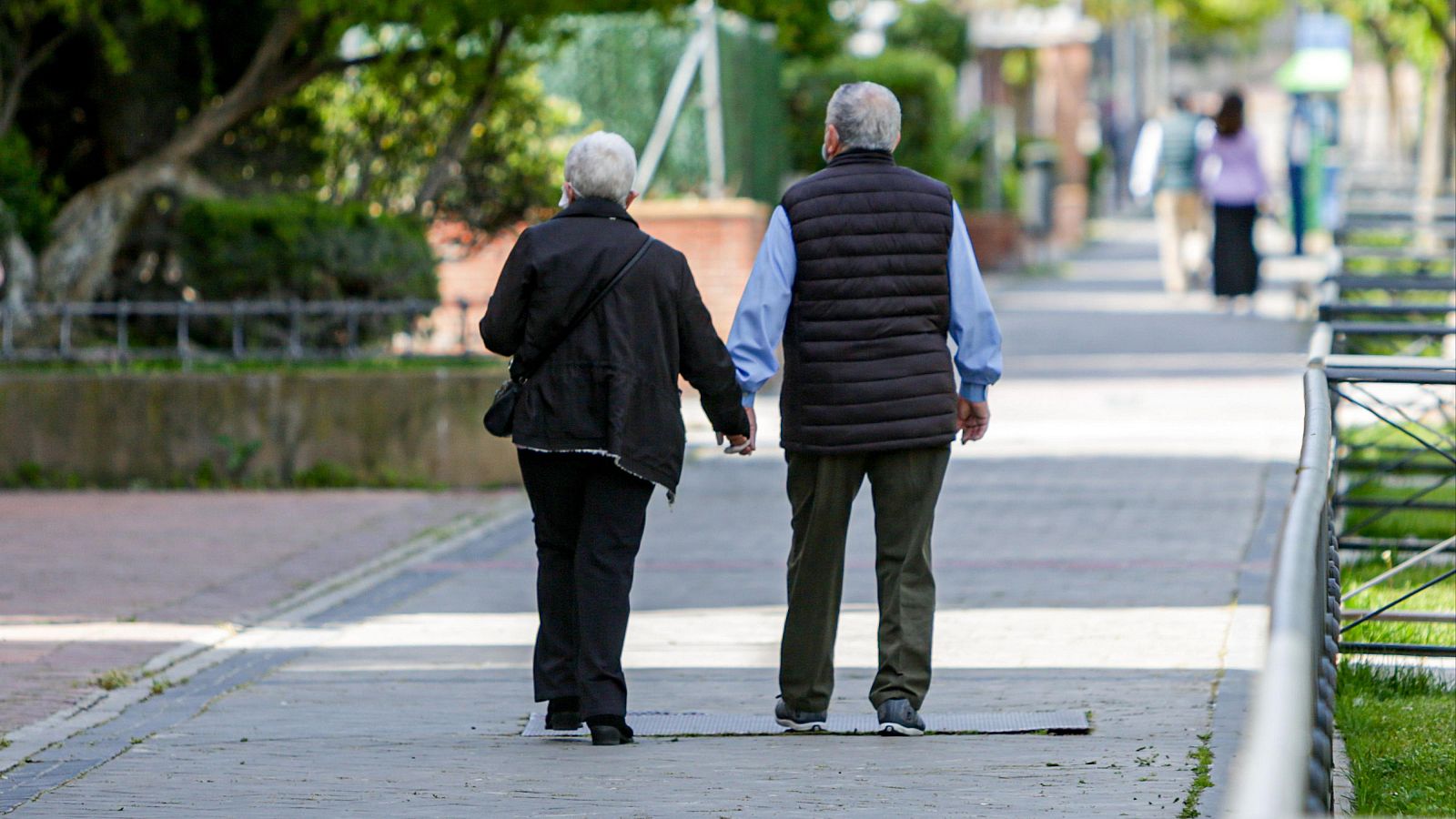 Pareja de ancianos paseando cogidos de la mano por un parque, vestidos con ropa abrigada.  Se alejan de la cámara, mostrando solo sus espaldas.