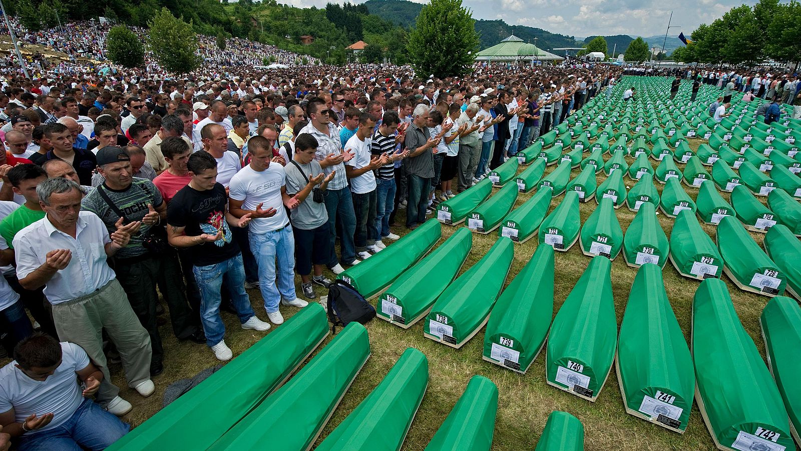 En un campo, se observa una ceremonia conmemorativa con ataúdes cubiertos y una multitud de personas orando, bajo un cielo nublado.