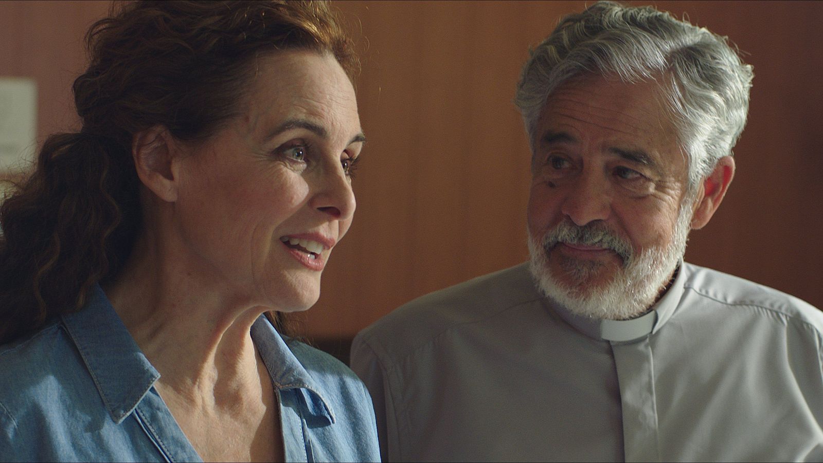 En primer plano, Silvia Marsó sonríe con una camisa vaquera, mientras que Carlos Iglesias, con alzacuello, la observa. El fondo difuminado es de color marrón claro.