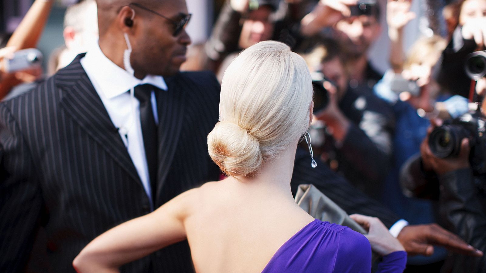 Una mujer con un vestido morado y un moño es escoltada por un hombre con gafas de sol y traje a rayas, mientras una multitud de fotógrafos intenta capturar imágenes.