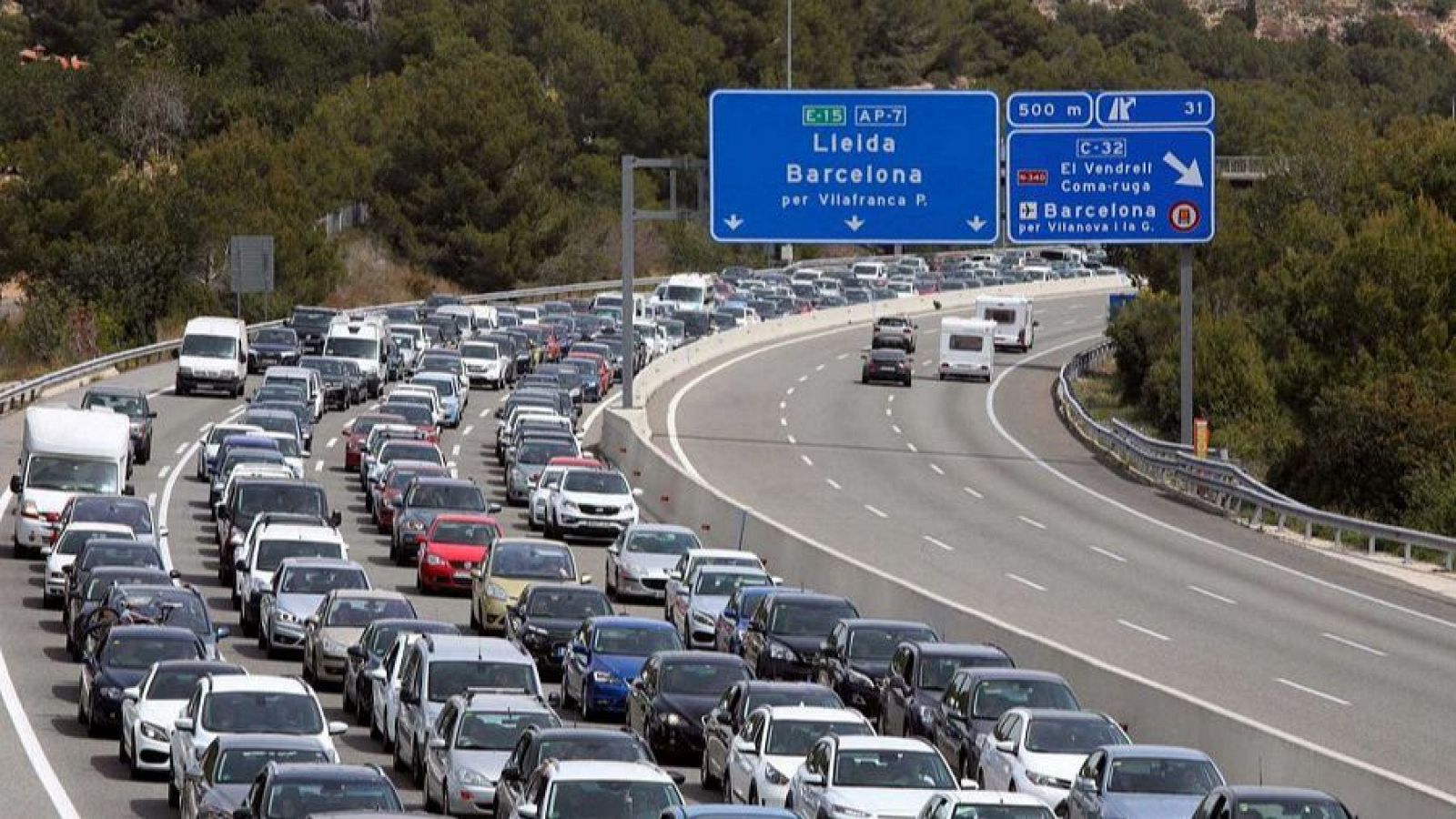 Atasco en autopista con coches de diferentes tipos. Señales indican direcciones a Lleida y Barcelona. Día soleado.