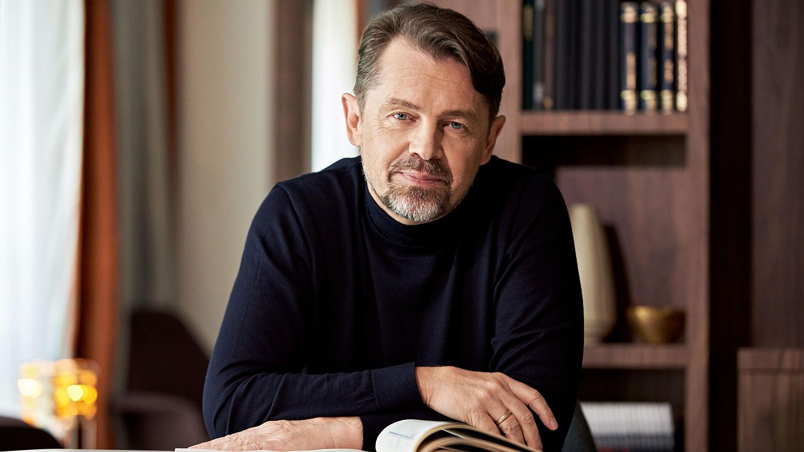 Un hombre de mediana edad, con cabello castaño peinado hacia atrás y barba incipiente, sonríe levemente mientras está sentado frente a un libro abierto. Viste un jersey de cuello alto azul oscuro.