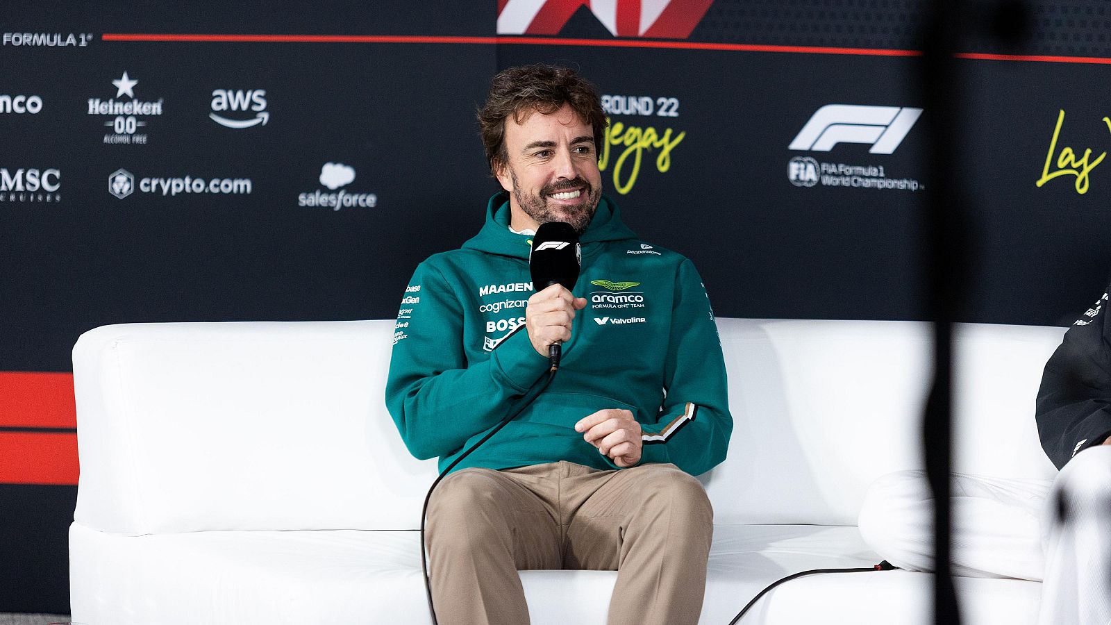 El piloto de Fórmula 1, Fernando Alonso, está sentado en un sofá, con una sudadera verde y un micrófono, durante una rueda de prensa con un fondo negro lleno de logotipos.