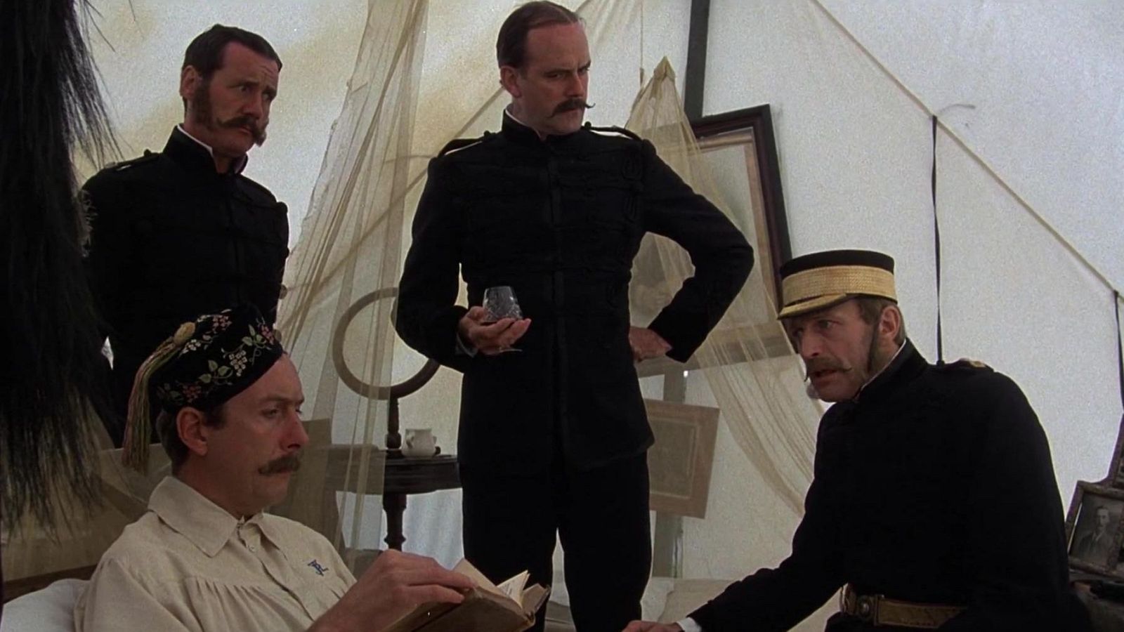 Cuatro hombres, tres con uniformes militares oscuros y bigotes, se encuentran en un espacio interior. Uno sostiene una copa y otro lee un libro con un fez. Hay una mesa con una taza y otros objetos.