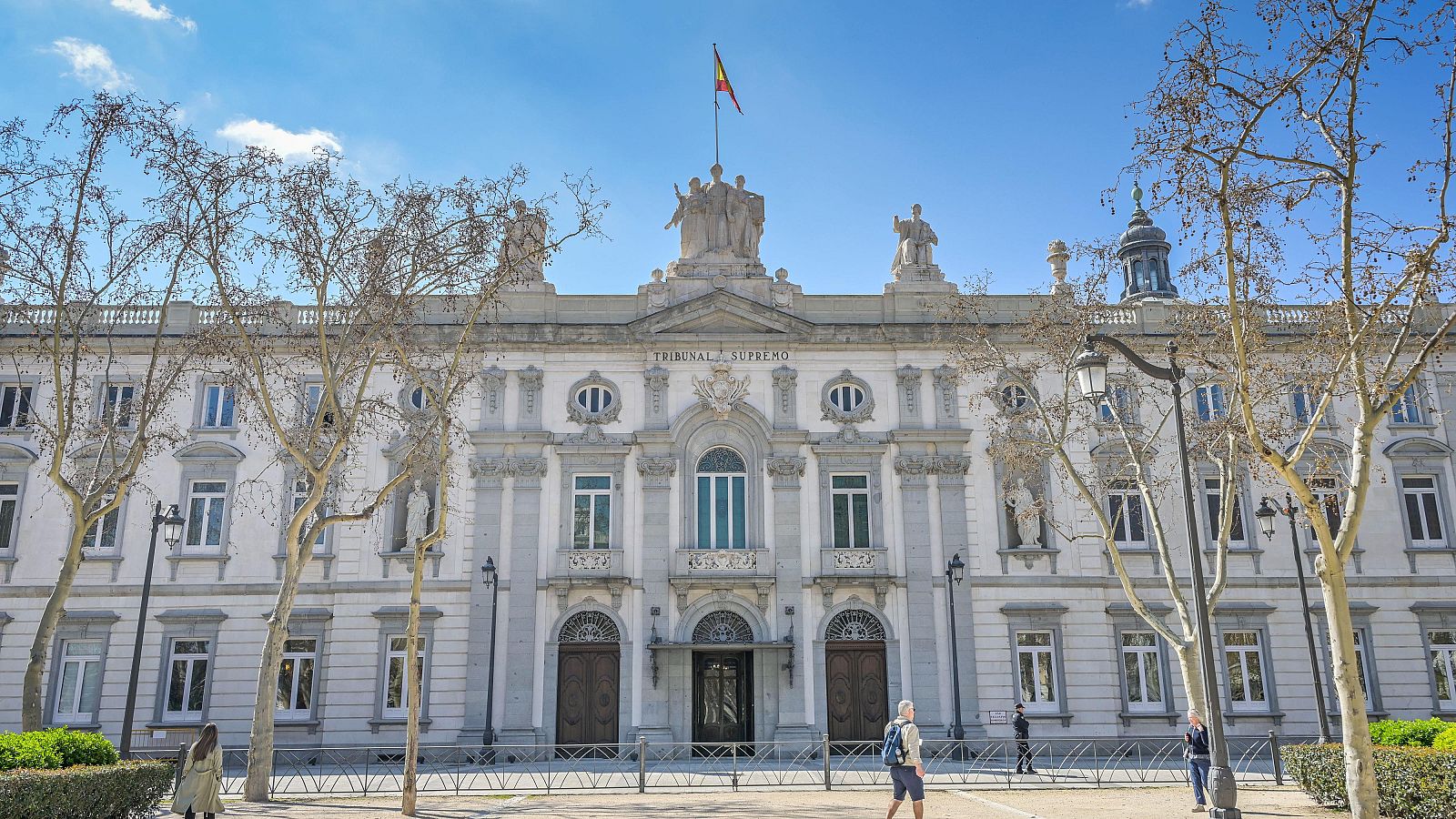 Se observa la fachada de un edificio de piedra blanca con múltiples ventanas y una inscripción en la parte superior. Frente al edificio, hay una plaza con árboles y farolas, con algunas personas caminando.