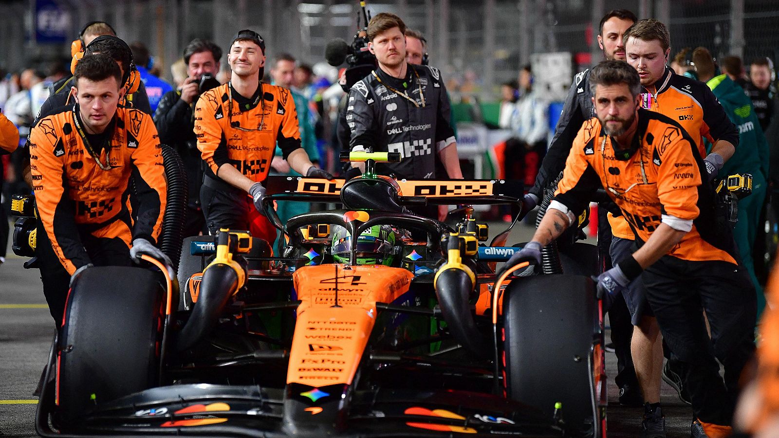 Un equipo de mecánicos de McLaren trabaja en el coche de Fórmula 1, con el número 4, durante una carrera. El monoplaza, de color naranja y negro, se encuentra en el pit lane, con otros equipos y público al fondo.