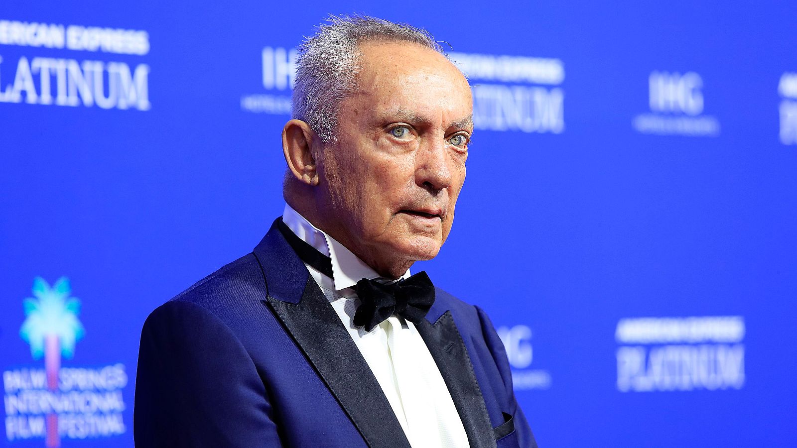 Udo Kier, con cabello canoso y expresión serena, luce un esmoquin azul oscuro con detalles en negro. El fondo azul muestra logotipos y textos promocionales.