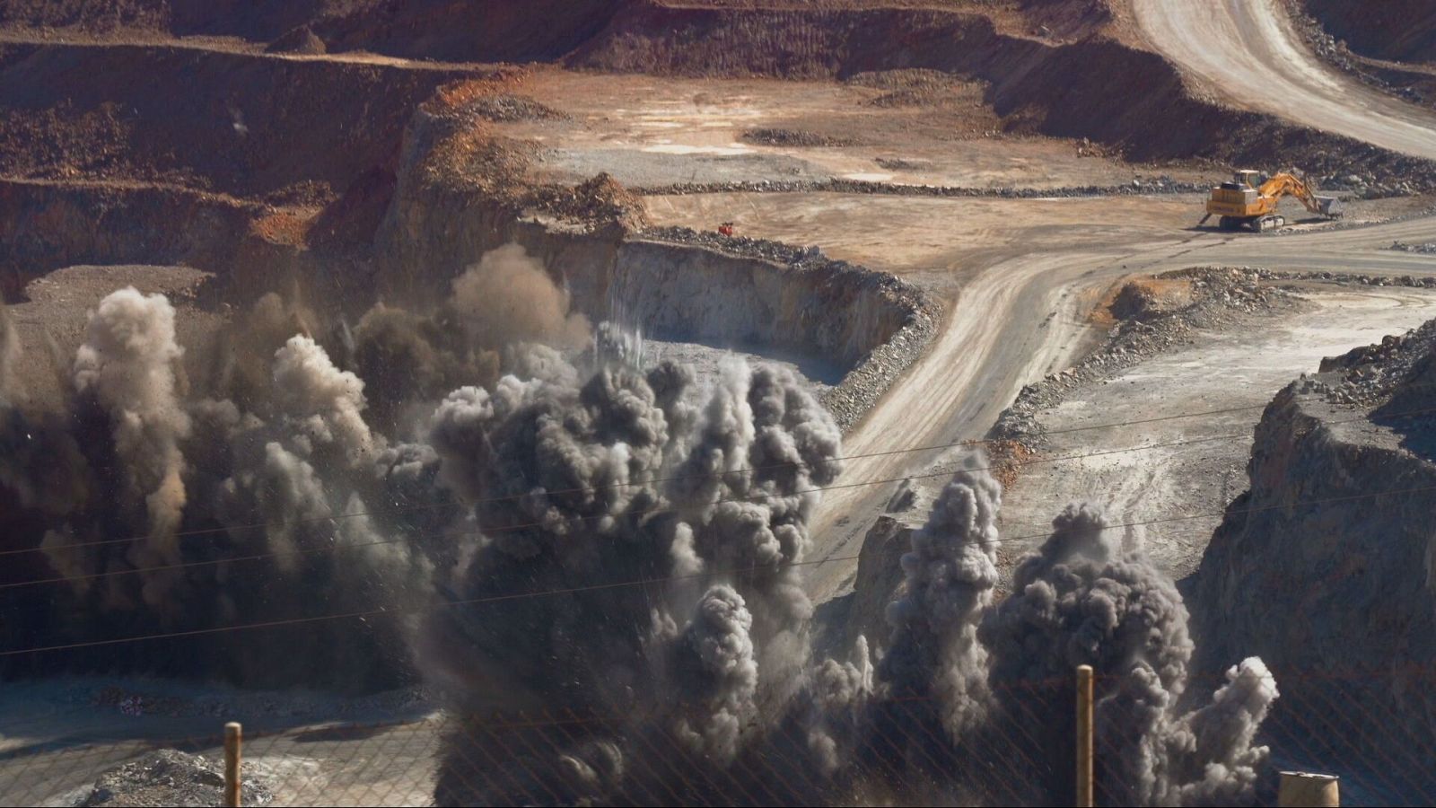 Una detonación controlada en una mina a cielo abierto produce una gran columna de humo. Una excavadora amarilla opera en la superficie, con caminos de tierra que cruzan el terreno irregular y rojizo.