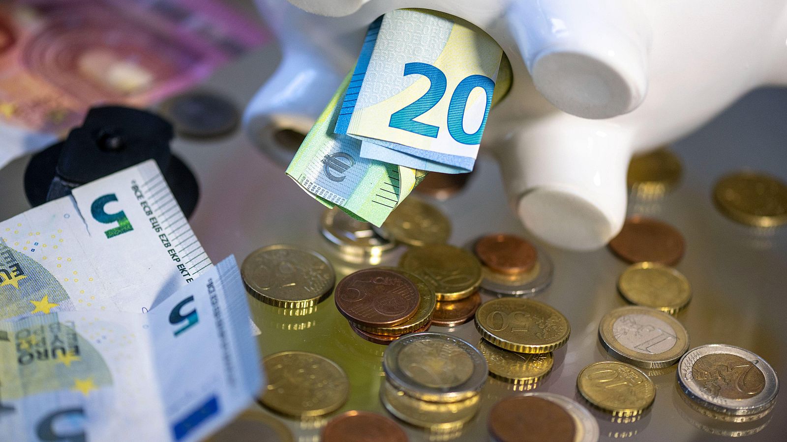 En la imagen, una hucha con forma de cerdo, de color blanco, está llena de monedas y billetes. Se observa un billete de 20 euros enrollado y otro parcialmente visible, junto con monedas de diferentes valores.
