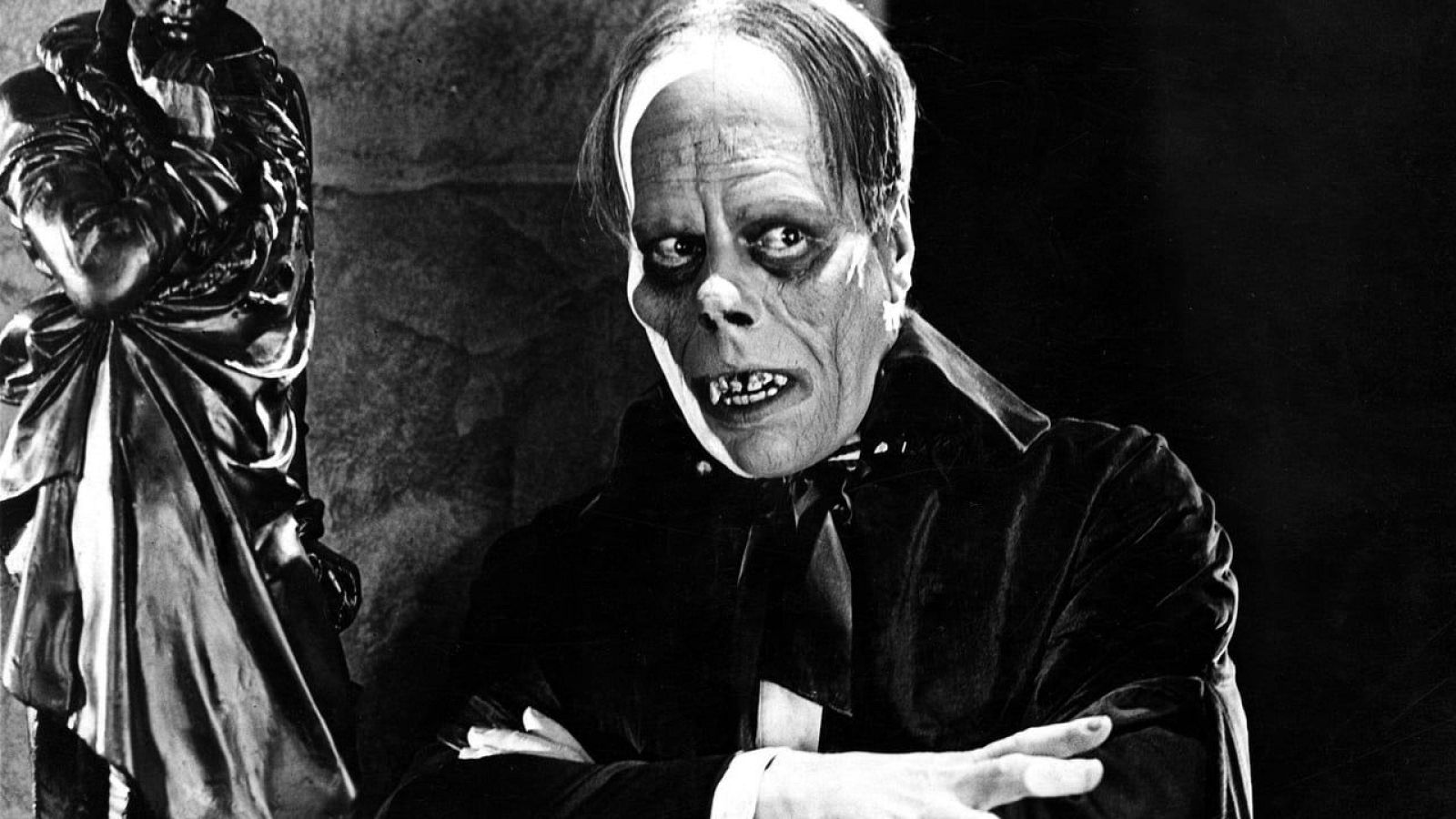 En la fotografía en blanco y negro, un hombre con maquillaje pálido y deformado, con los brazos cruzados, personifica a un personaje con ojos hundidos y dentadura visible, vestido con traje oscuro y corbata negra, en un ambiente sombrío.