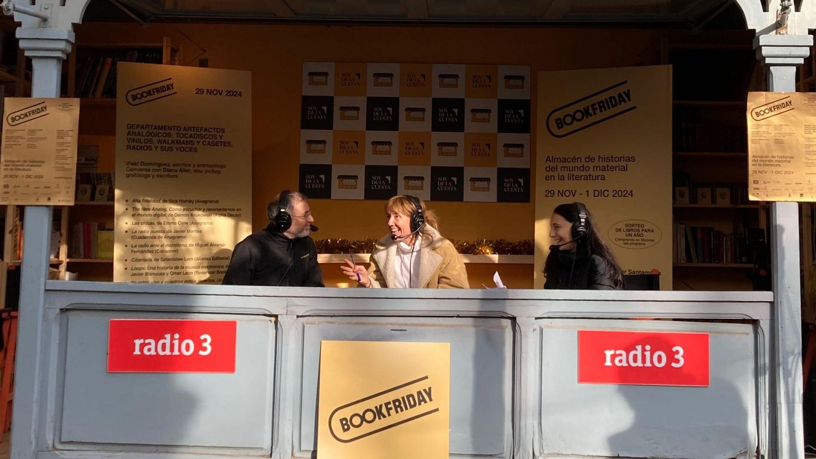 En el estudio de radio, tres personas con auriculares y micrófonos participan en un evento. Detrás de ellos, un panel y carteles promocionan el evento, con estanterías llenas de libros en el fondo.