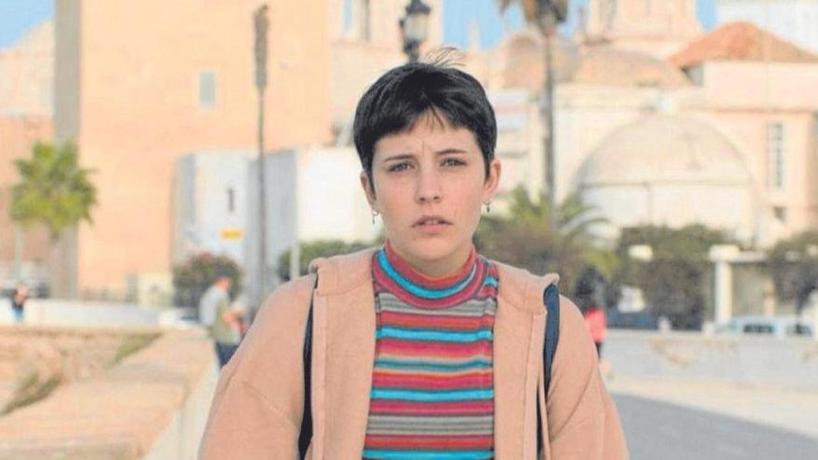 Una joven con cabello oscuro y camiseta de rayas, posiblemente en un fotograma de una película, posa en un entorno exterior con edificios y palmeras, llevando una mochila.