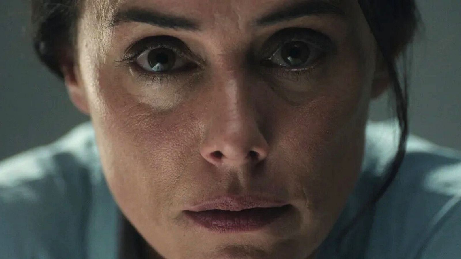 Primer plano del rostro de una mujer con ojos oscuros y mirada penetrante, con la piel de textura natural y labios finos, en un ambiente de iluminación tenue y dramática.