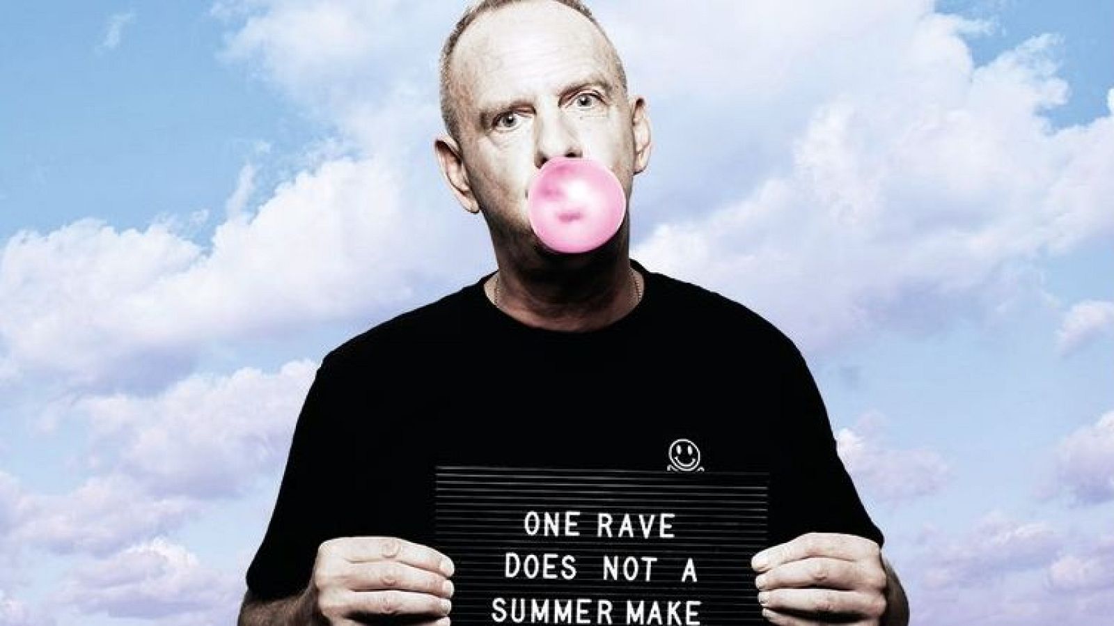 Un hombre con una expresión seria, con chicle en la boca, sostiene un cartel con texto en letras blancas, vistiendo una camiseta negra con un logo.