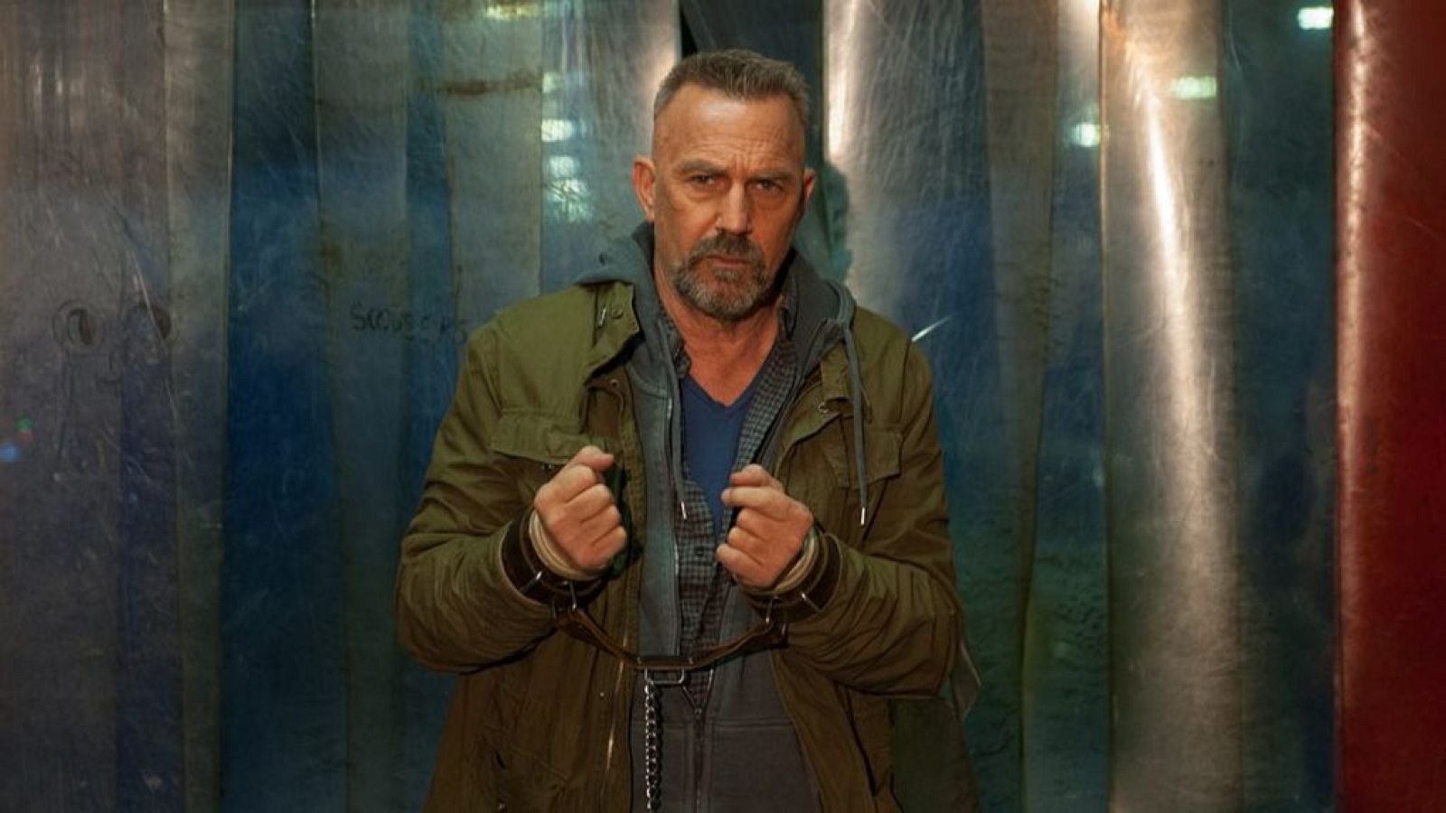Kevin Costner, como Jerico Stewart, aparece con el pelo canoso y barba gris, vistiendo una chaqueta verde oliva y camisa a cuadros, con las manos esposadas frente a una pared metálica.