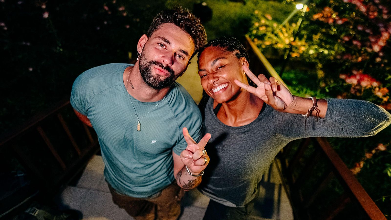Dos personas sonríen y posan juntas al aire libre. Un hombre con barba y camiseta azul hace el signo de la paz junto a una mujer con trenzas y camiseta gris.