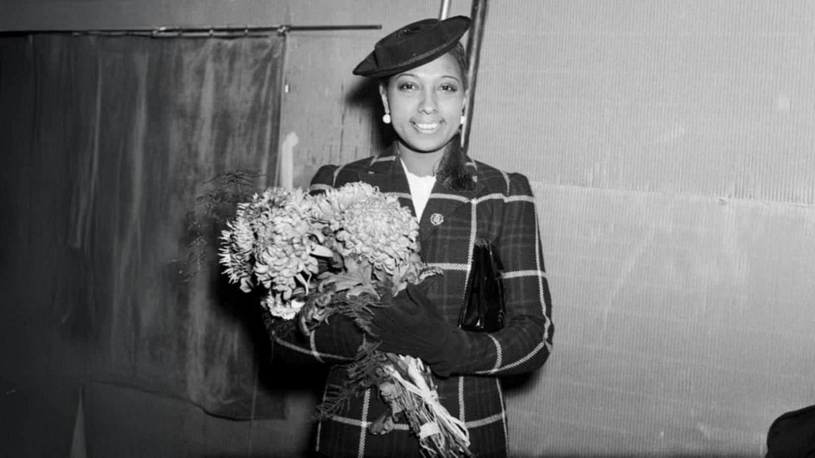 Josephine Baker, fotografiada en 1938, sonríe a la cámara, luciendo un sombrero oscuro, pendientes de perlas y un abrigo a cuadros, mientras sostiene un ramo de flores.