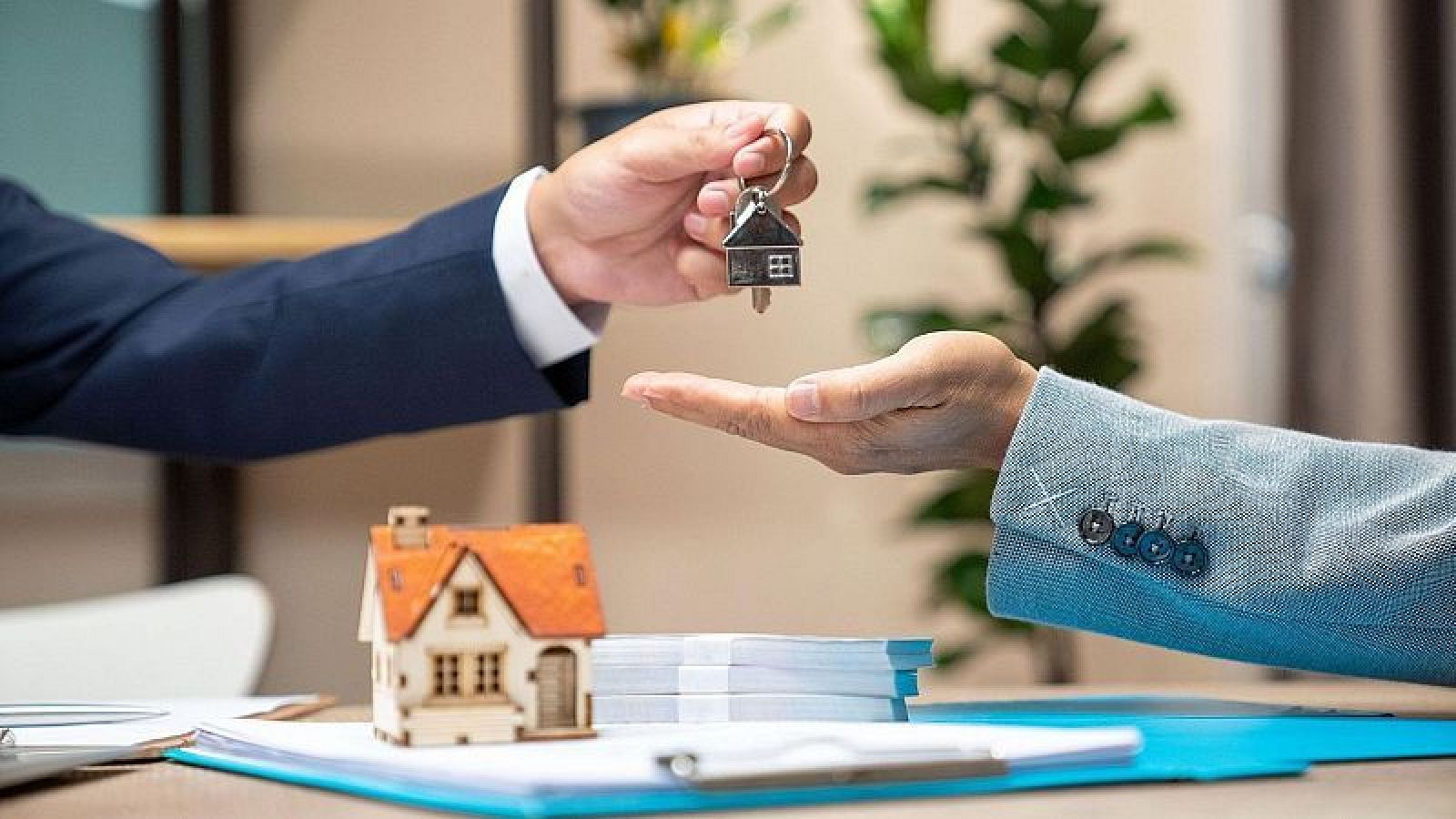 Una mano con chaqueta azul marino entrega llaves a otra mano con chaqueta azul claro, con una maqueta de casa y dinero sobre la mesa, posiblemente en una transacción de compraventa.
