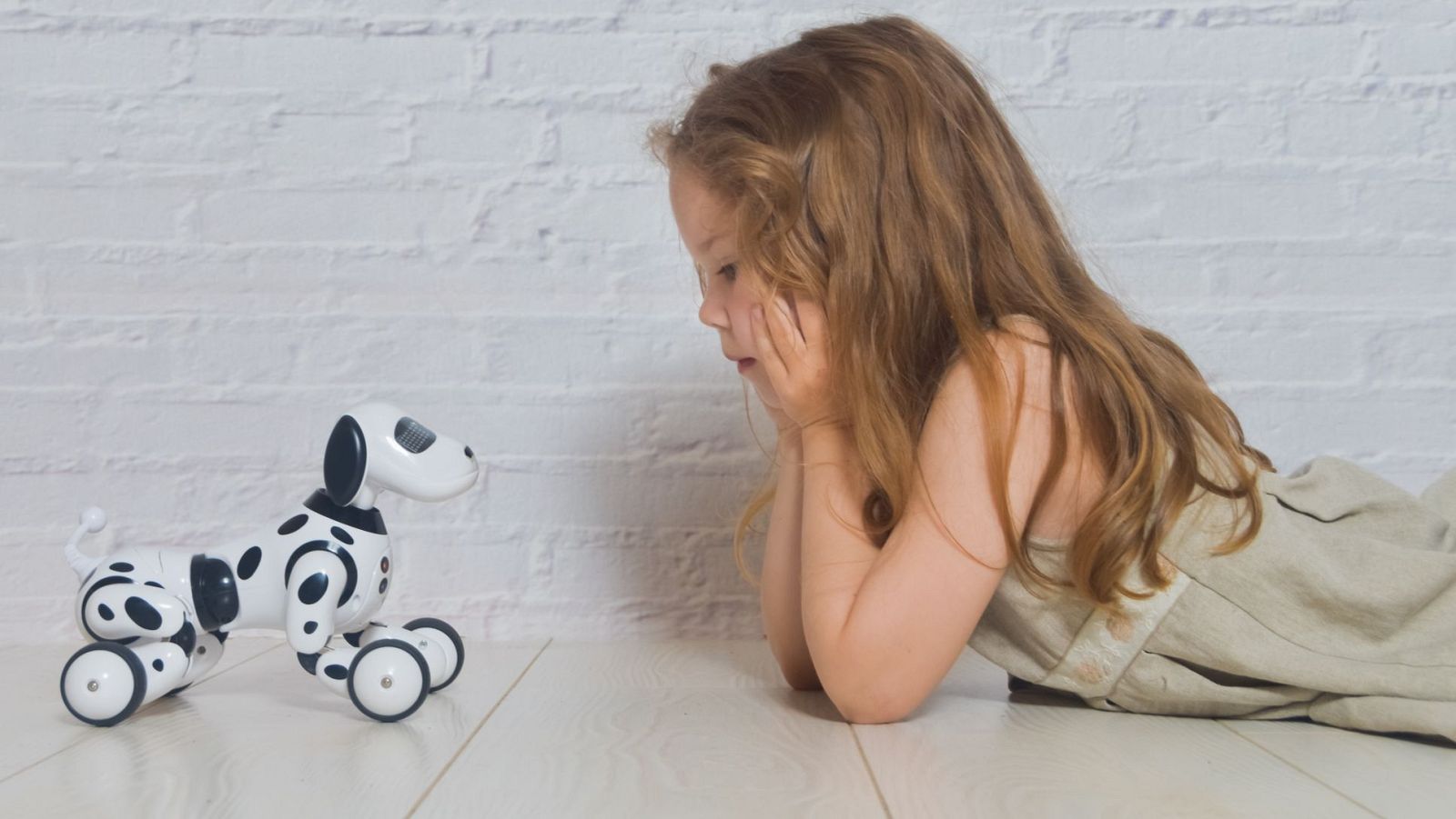 Una niña pequeña, con vestido claro, interactúa con un juguete robótico con forma de perro dálmata en un suelo de madera. El juguete, con ruedas y manchas negras, parece ser un dispositivo de compañía.