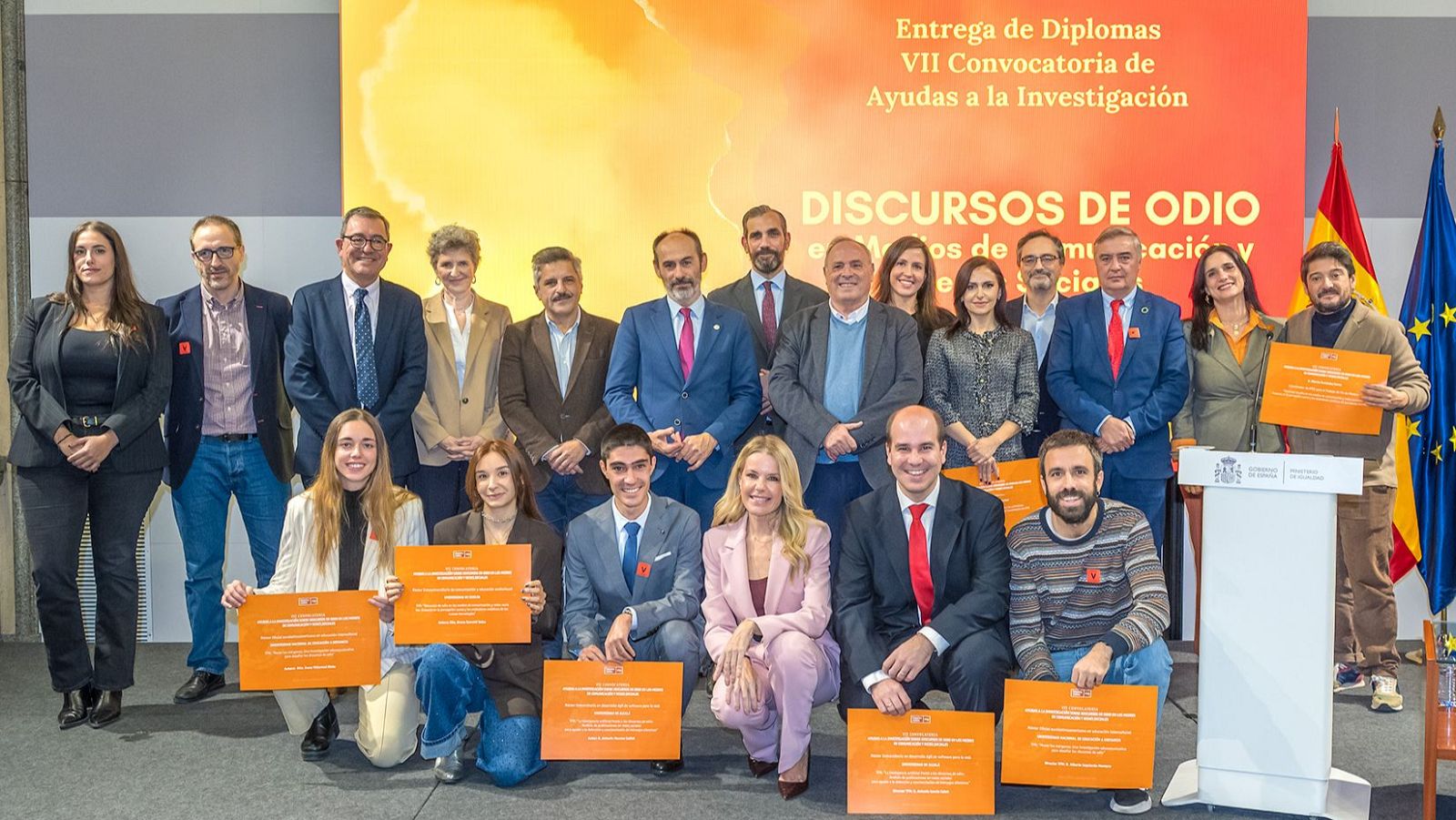 Un grupo de personas posa para una foto en un evento de entrega de diplomas. Se observa a una presentadora y actriz, junto a asistentes que sostienen diplomas. El fondo muestra el título del evento y banderas.