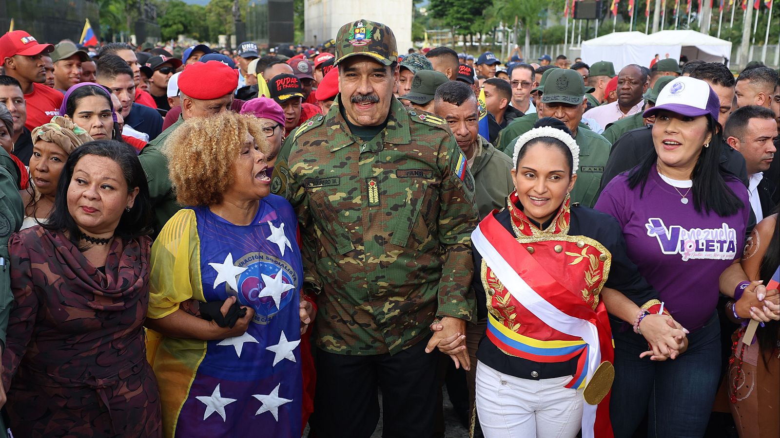 Nicolás Maduro, vestido con uniforme militar, se encuentra rodeado de personas, incluyendo dos mujeres que portan elementos con los colores de la bandera venezolana, en un acto público donde se observan banderas ondeando.