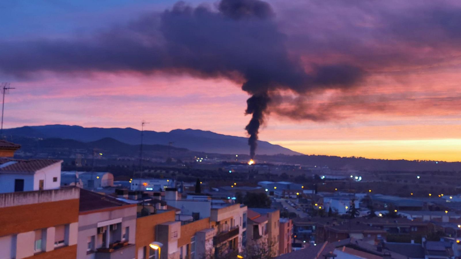 Una imatge captura un incendi a distància, amb una columna de fum negre que s'eleva cap al cel tenyit de colors càlids. En primer pla, es distingeixen teulades d'edificis residencials, mentre que al fons s'alcen muntanyes.