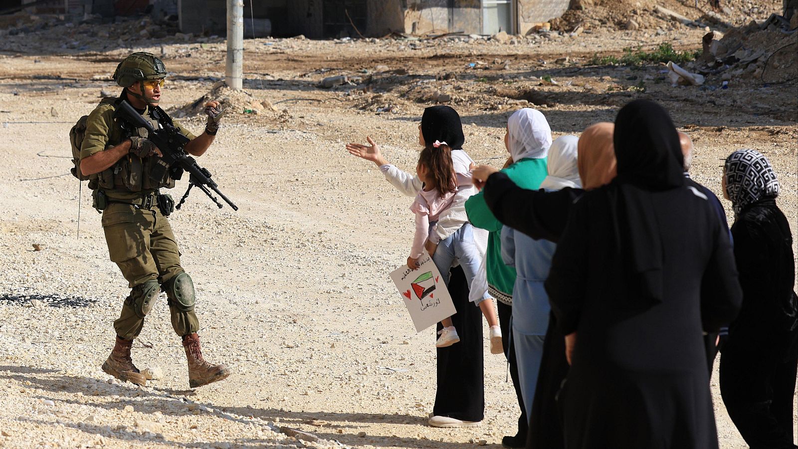 Un soldado israelí impide el paso a un grupo de mujeres y niños. Una niña porta un cartel con símbolos y escritura. El fondo muestra un terreno desolado y construcciones.