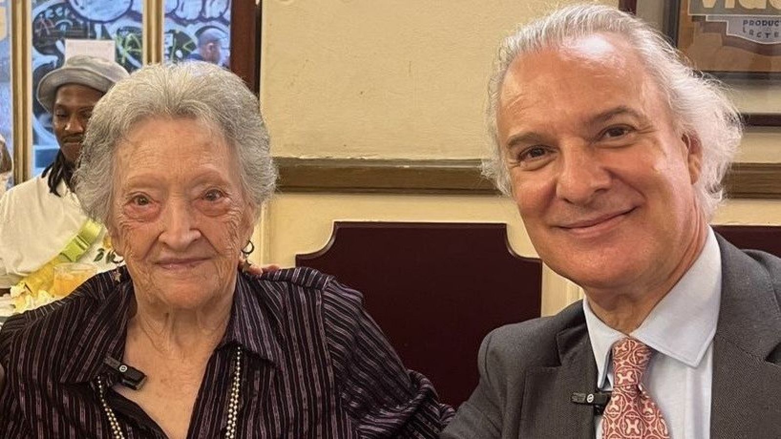 Dos personas, una mujer mayor con blusa a rayas y un hombre de mediana edad con traje gris, posan con micrófonos. Se aprecia un cartel con la leyenda "PRODUCTOS LACTEOS".