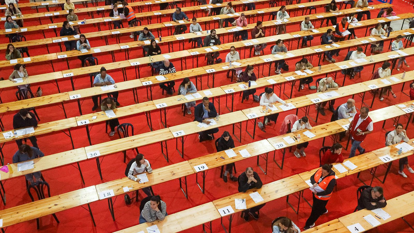 Una sala espaciosa, con mesas numeradas y dispuestas en filas sobre una alfombra roja, alberga un examen. Los participantes están concentrados en sus documentos, supervisados por una persona con chaleco reflectante.