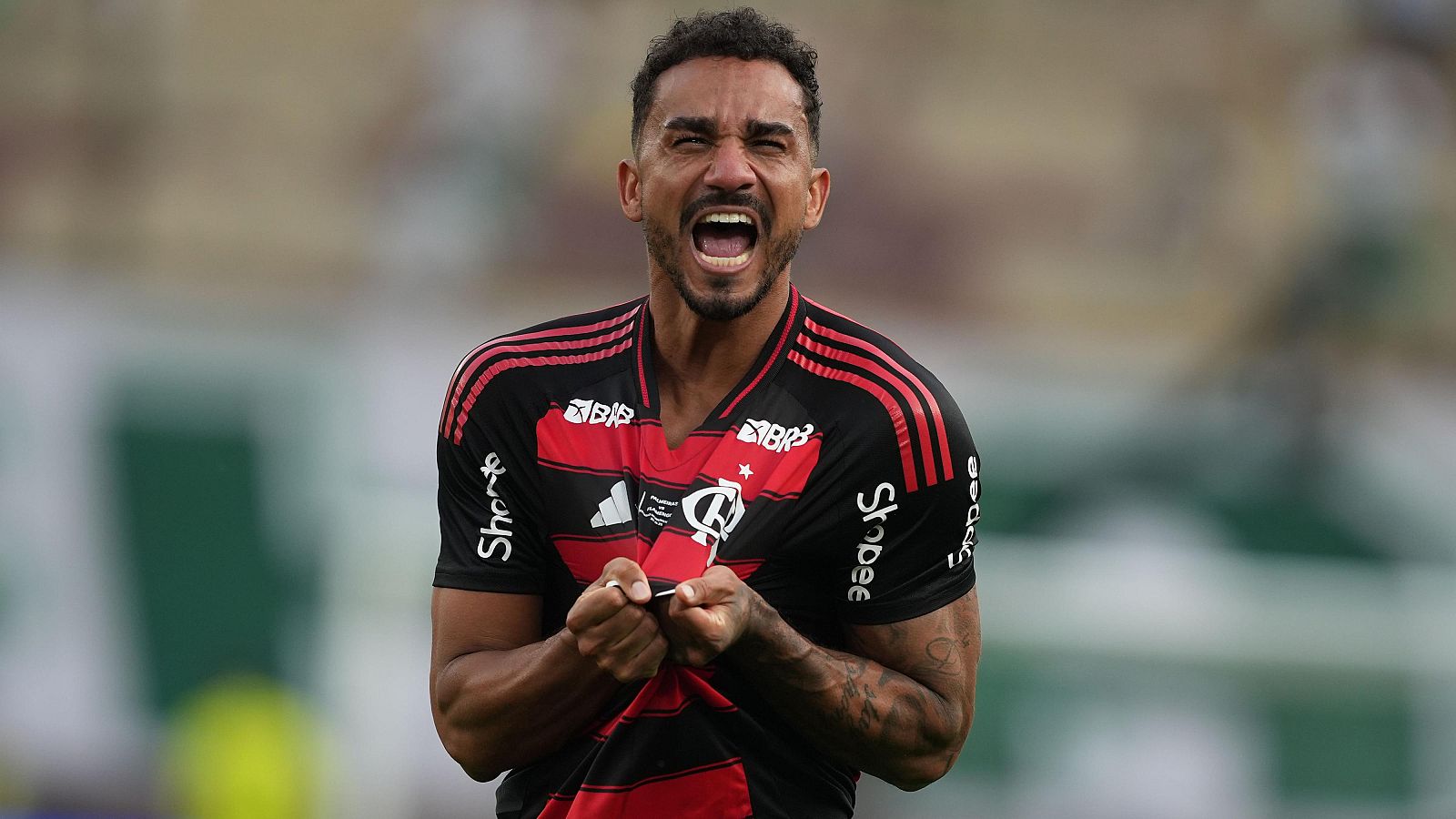 Un jugador del Flamengo, con la camiseta a rayas rojas y negras, celebra con los puños cerrados y una expresión de alegría en un campo de fútbol, mostrando el escudo del equipo y logos de patrocinadores.
