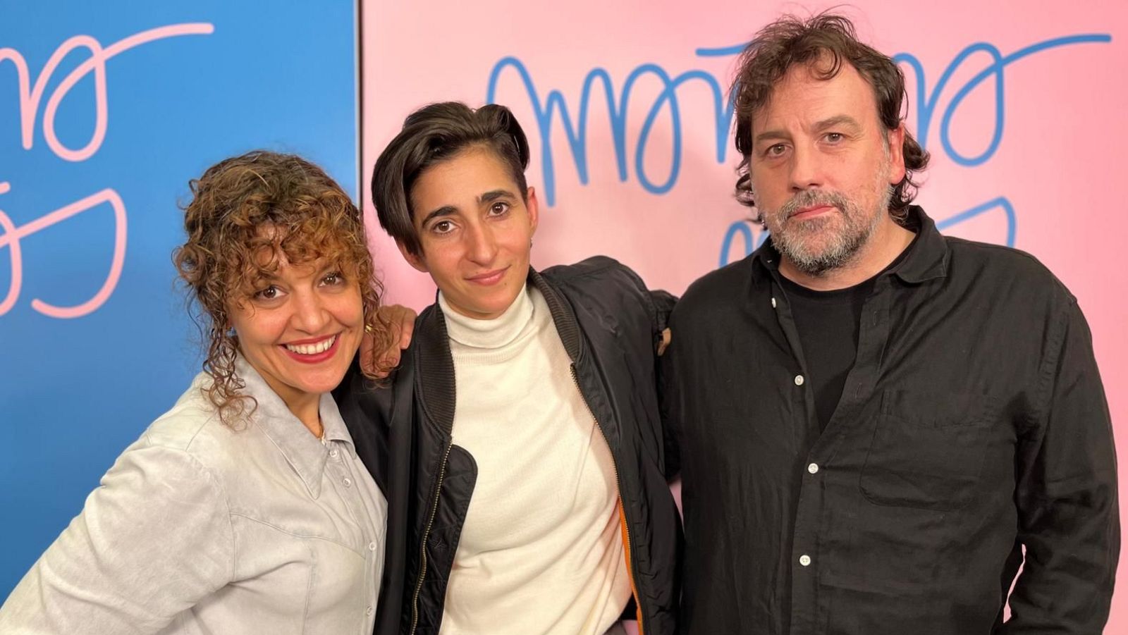 Tres personas posan frente a una pared dividida en dos colores con texto. Una mujer con camisa clara sonríe a la izquierda, otra con jersey blanco y chaqueta negra está en el centro, y un hombre con camisa negra y barba se encuentra a la derecha.