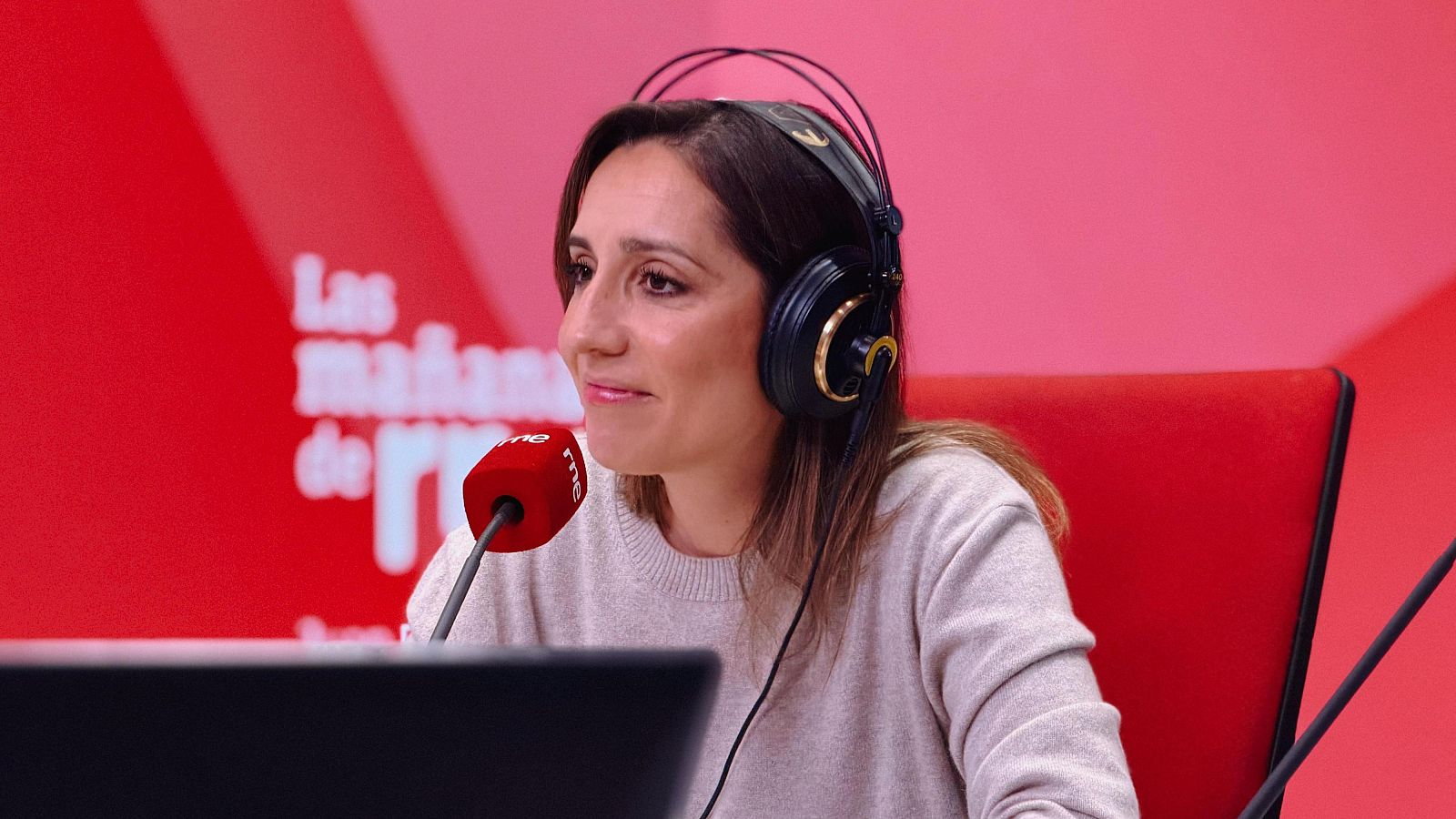 Ángela Banzas, con auriculares y suéter crema, se encuentra frente a un micrófono rojo con el logo de una emisora, en un estudio con fondo rojo, posiblemente para una grabación.