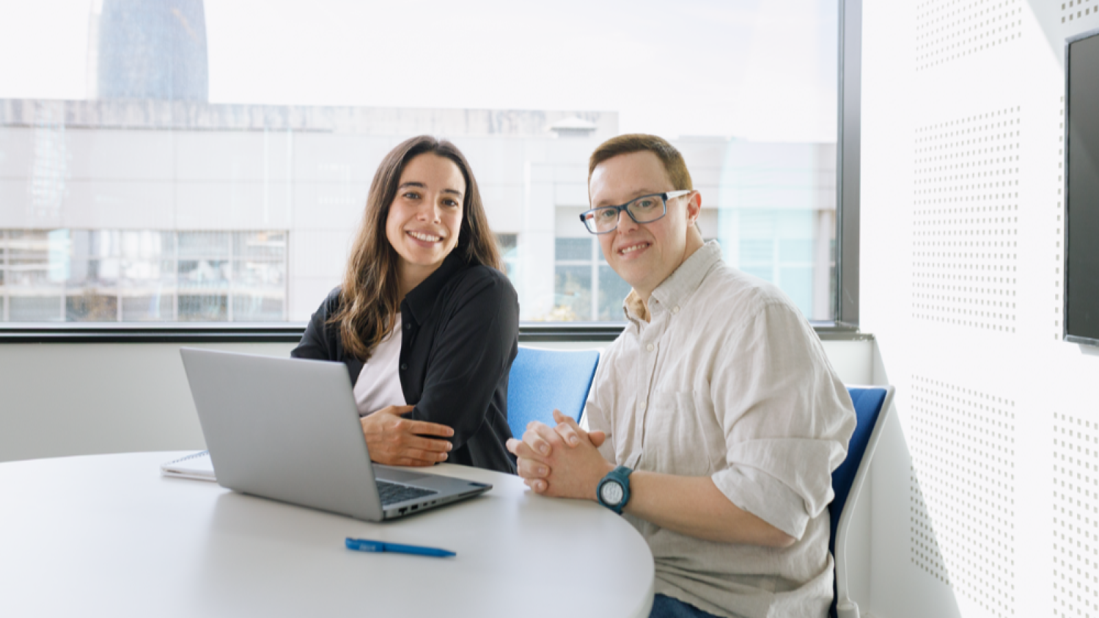Dos personas sonríen a la cámara en una oficina: una mujer frente a un portátil y un hombre con gafas y reloj azul. Hay un bolígrafo azul sobre la mesa y una ventana con un edificio al fondo.