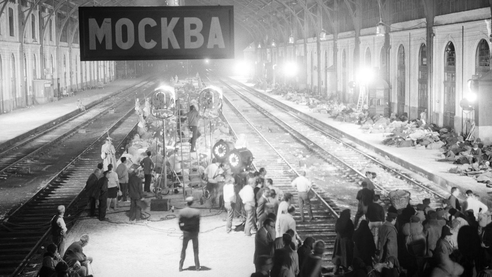 En una estación, un equipo de filmación trabaja en una escena con extras y un letrero en ruso. La iluminación intensa y la arquitectura clásica crean el ambiente deseado.