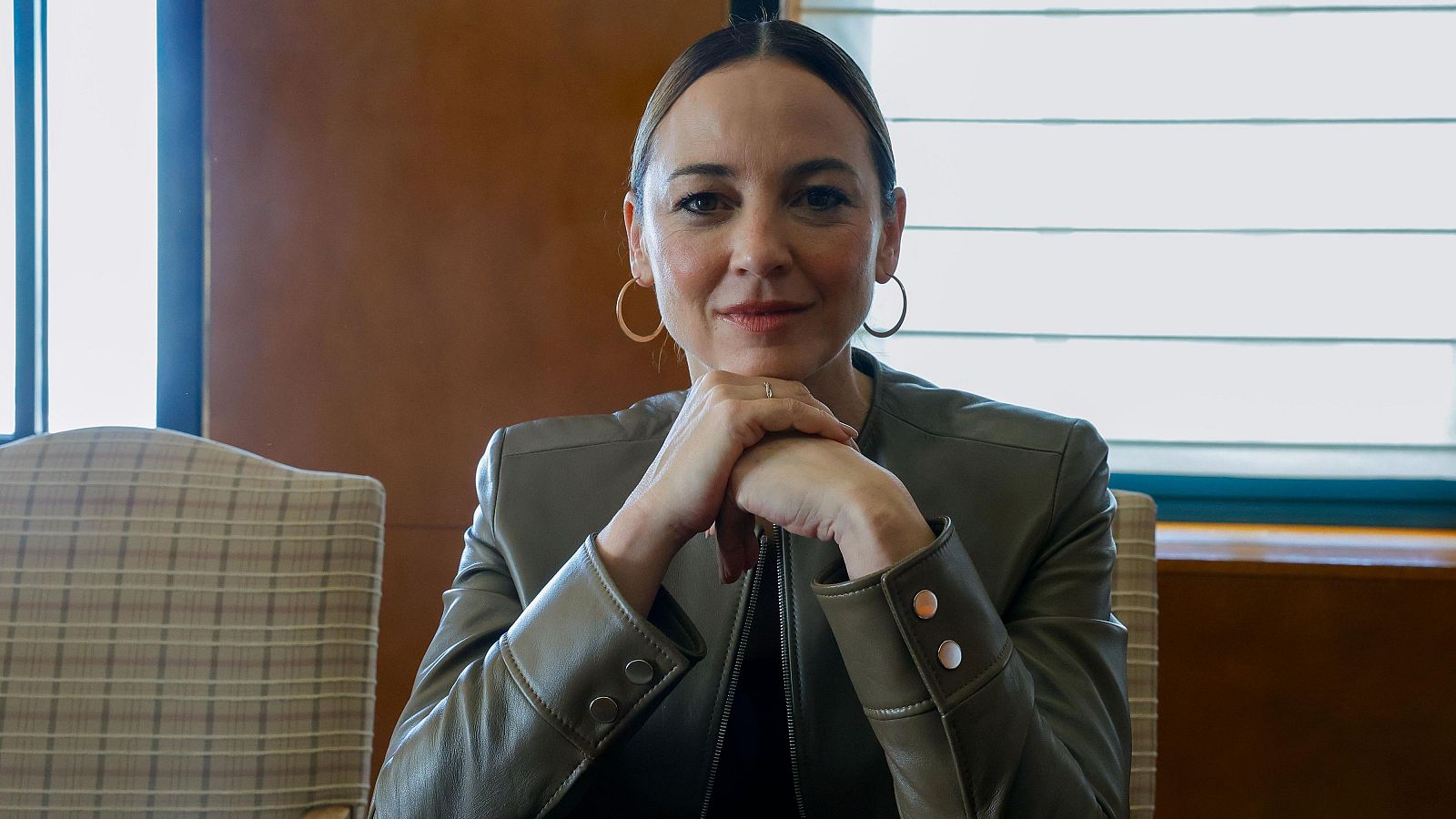 Una mujer con chaqueta de cuero verde oliva y pendientes dorados, con el pelo recogido, posa con las manos entrelazadas y apoyadas en la barbilla, mostrando una expresión serena. El fondo sugiere una habitación con luz natural.