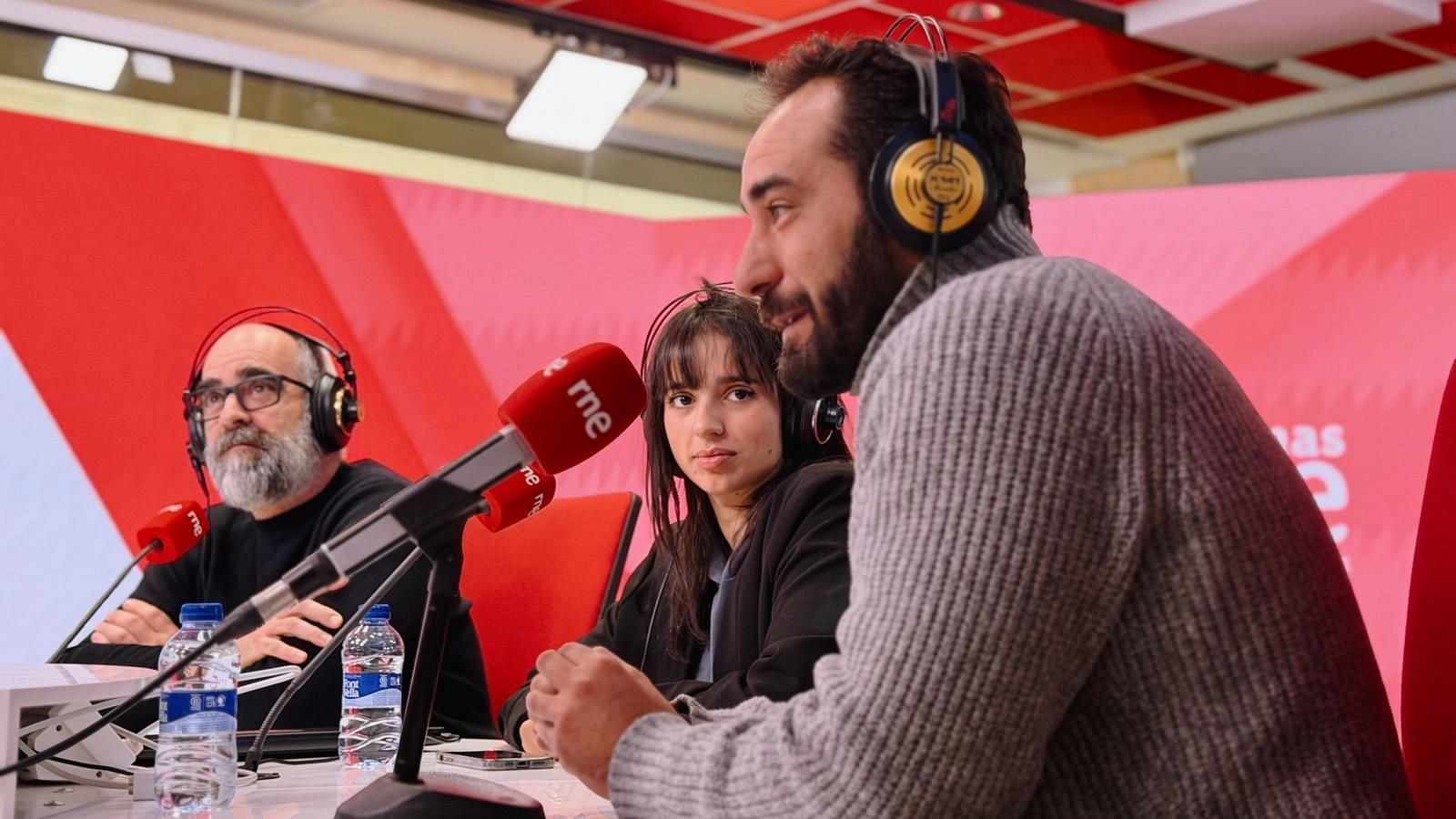 Tres personas, dos hombres y una mujer, se encuentran en un estudio de radio frente a micrófonos. Los hombres llevan auriculares y barba, mientras que la mujer tiene el pelo oscuro y flequillo. Hay botellas de agua sobre la mesa.