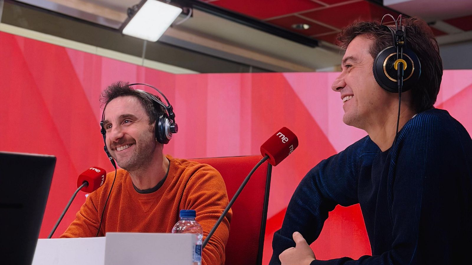 Dos hombres en un estudio de radio, con auriculares y micrófonos, se preparan para una emisión. Uno viste un jersey naranja y el otro, uno azul oscuro, con una botella de agua sobre la mesa.