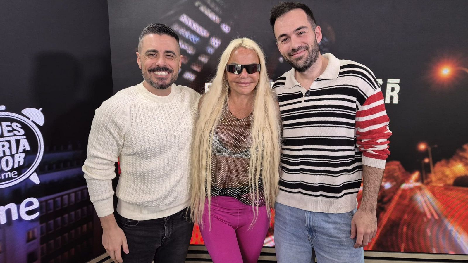Leticia Sabater, con gafas de sol y ropa llamativa, posa entre dos hombres. El fondo presenta un panel con el logo de un programa de radio y una imagen de una carretera.