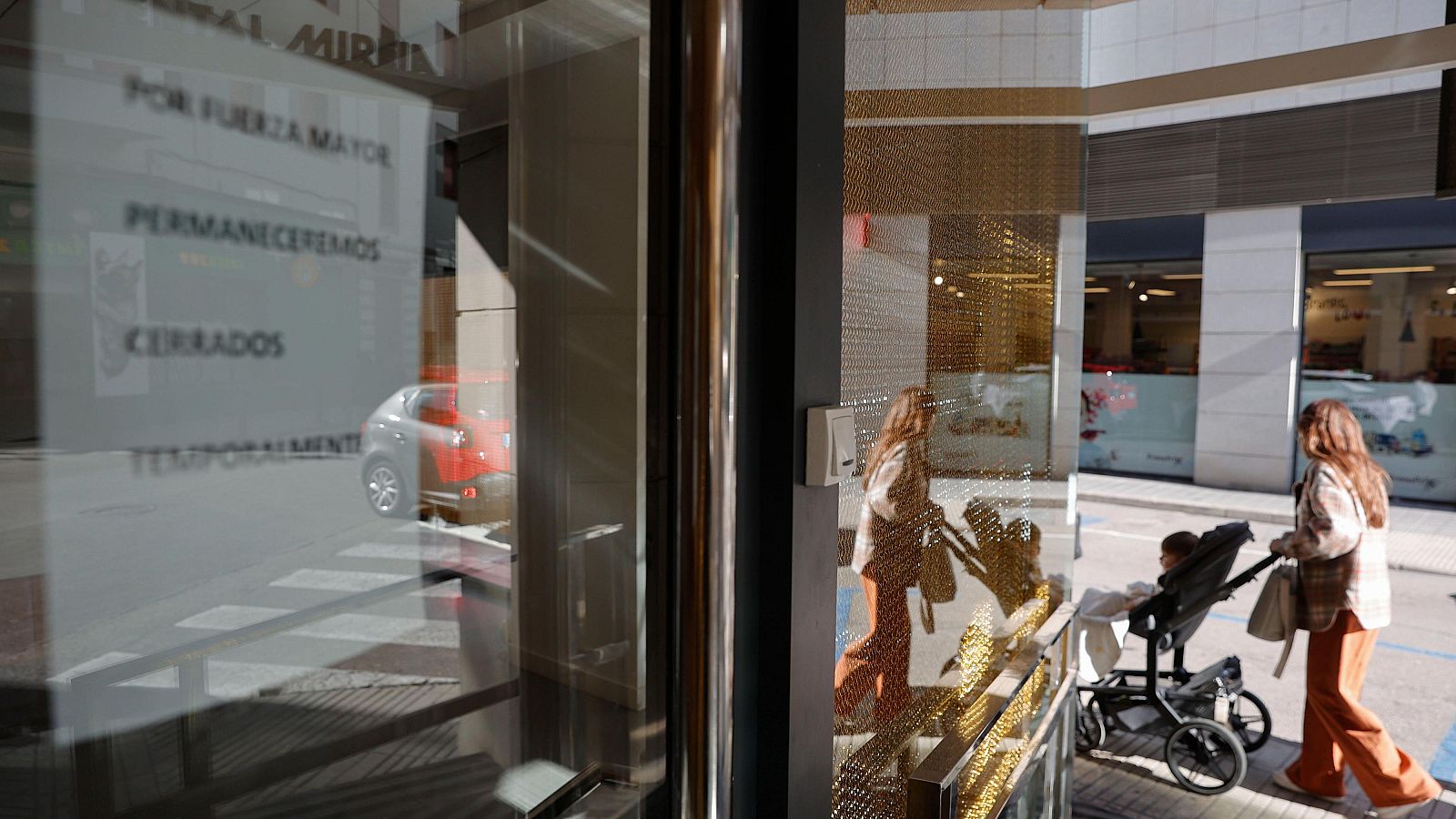 Fachada de una clínica dental con puerta de cristal y un letrero que anuncia un cierre temporal. En la calle frente a la clínica, se ve un coche rojo y una mujer con un carrito de bebé.