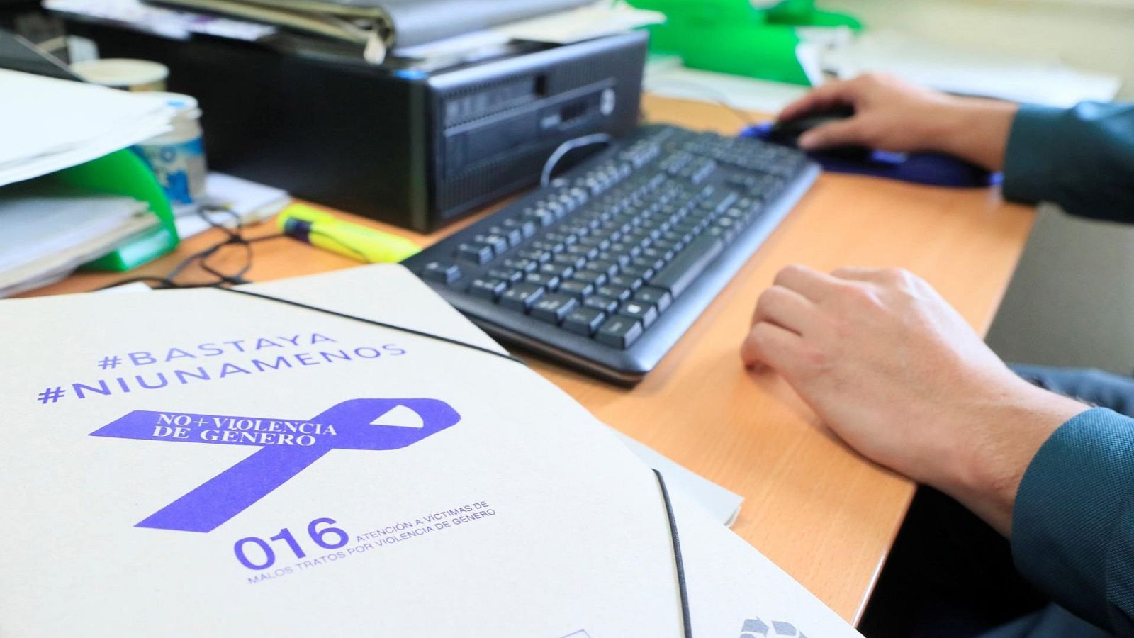 Un escritorio de trabajo muestra un ordenador en uso y una carpeta con información relevante sobre la lucha contra la violencia de género, incluyendo lemas y un número de teléfono de asistencia.