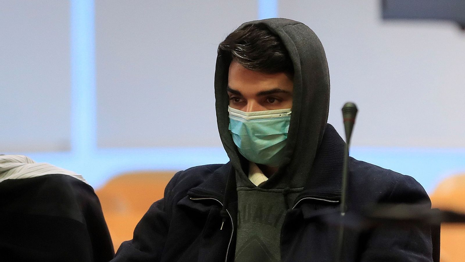 En la imagen se observa a un hombre joven, con mascarilla, sentado en un juzgado frente a un micrófono. Viste una sudadera con capucha y una chaqueta oscura. A su izquierda se distingue la presencia de otra persona.