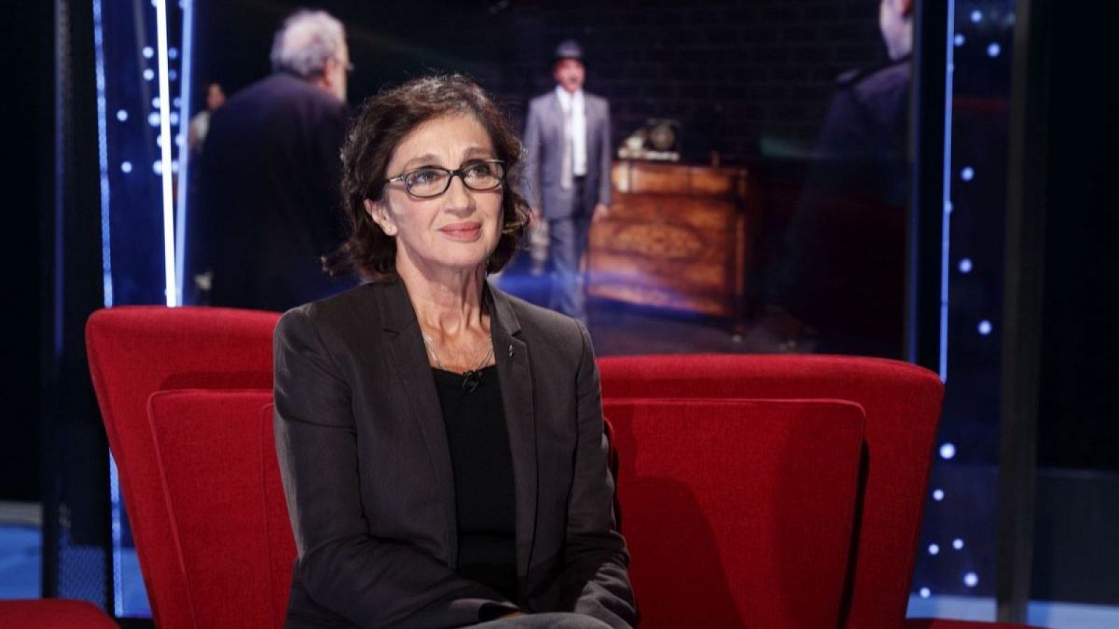 En la imagen, una mujer de mediana edad con gafas y cabello oscuro, vestida con chaqueta oscura y blusa negra, posa sentada en un sillón rojo frente a un decorado con una figura masculina y un mueble.