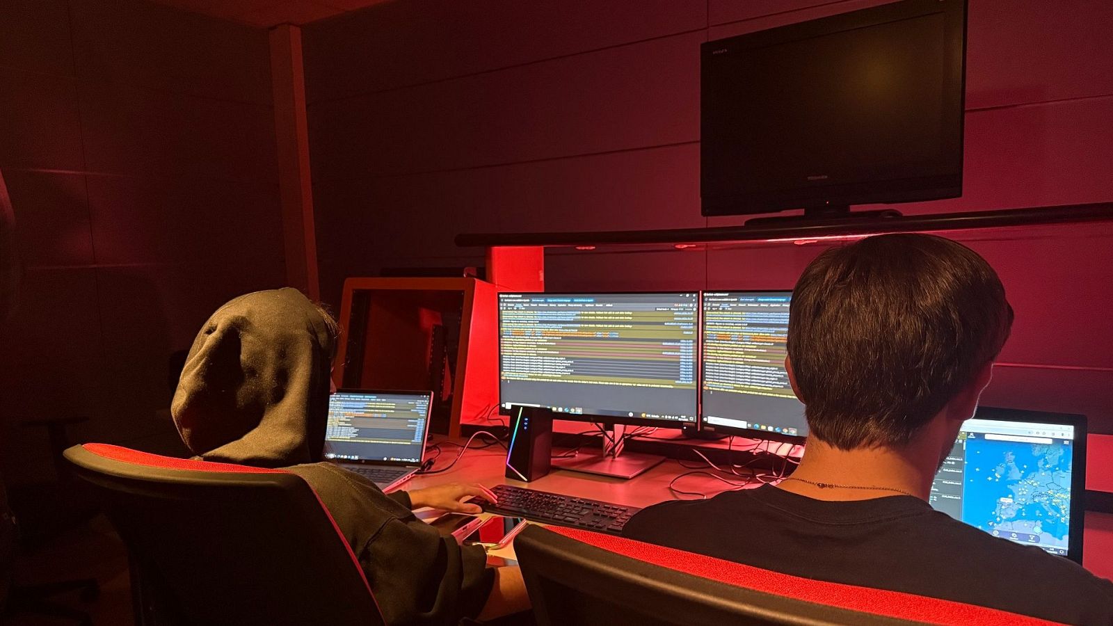 Dos individuos, uno con capucha y otro de espaldas, se encuentran en una habitación oscura con luces rojas, frente a varios monitores que muestran código y un mapa.